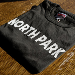 close up of details on a Black Peruvian Cotton tshirt with hand-finished details and "North Park" woven letters embroidered on the front in white. Features a logo patch on the bottom hem.